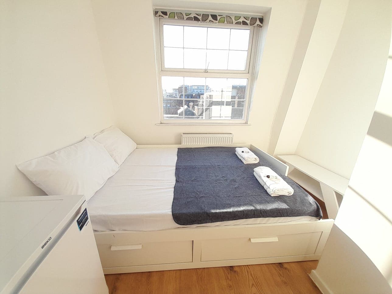 Bright bedroom with a white bed, grey throw, and towels next to a city-view window.