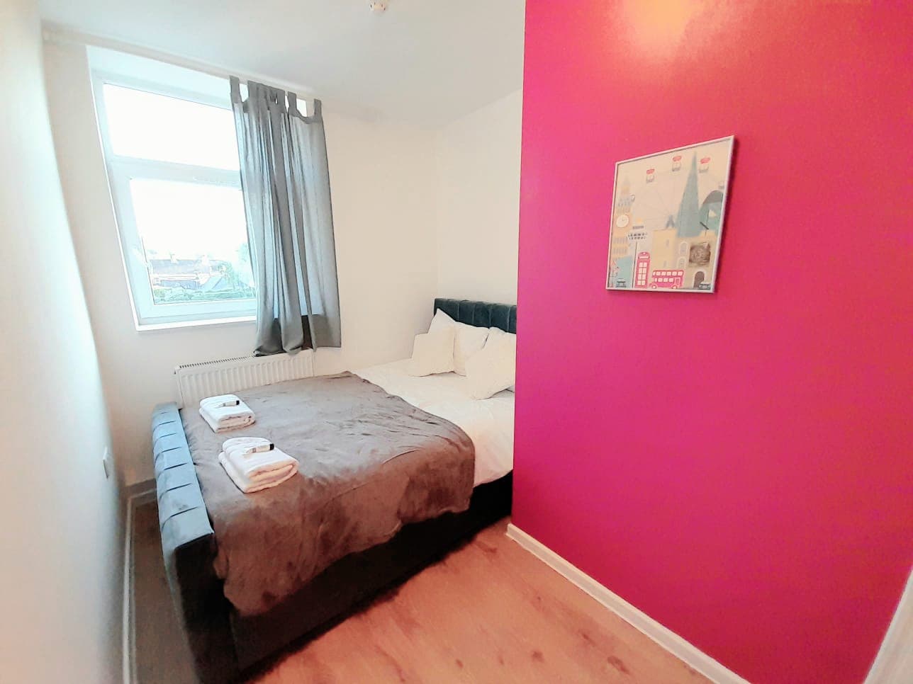 Small bedroom featuring a grey bed, white towels, a bright pink wall, and a window.